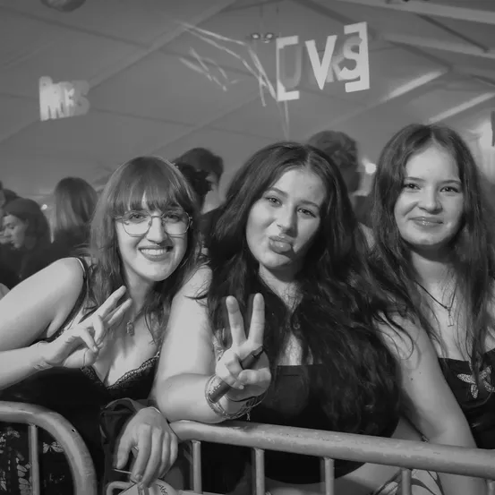 Drei Frauen bei einem Konzert machen Friedenszeichen, mit einem Schild oben, das GVG sagt, und einer Menge hinter ihnen.