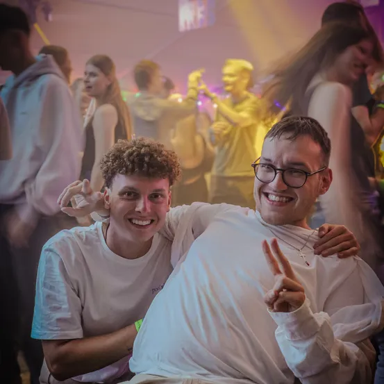 Zwei junge Männer lächeln und posieren für ein Foto in einer lebhaften Clubumgebung, während andere hinter ihnen tanzen.