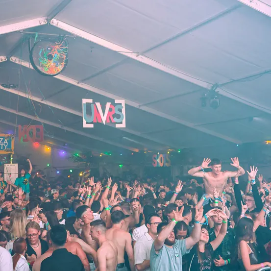 Ein überfüllter Indoor-Festival-Veranstaltungsort mit einer großen Menschenmenge. Ein Mann steht auf den Schultern einer Person in der Mitte. Ein farbenfrohes Schild hängt von der Decke.