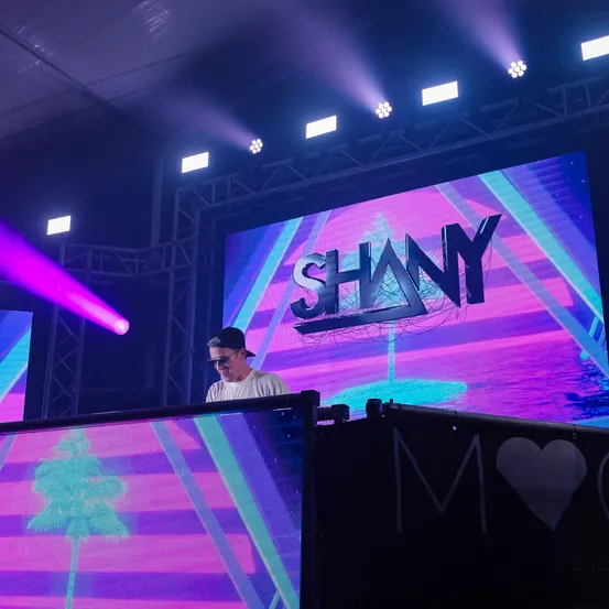 Ein Mann steht an einer DJ-Anlage mit einer großen Leinwand hinter ihm, auf der der Name 'SHANY' steht. Die Bühne ist mit leuchtenden lila und rosa Lichtern beleuchtet.