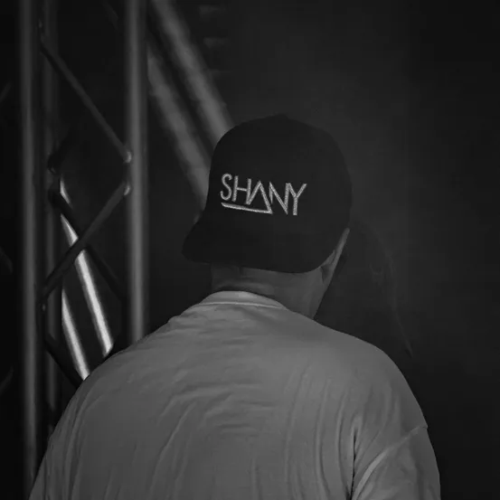 Ein Mann in einem weißen T-Shirt und einer schwarzen Kappe mit dem Wort 'SHANY' in weißen Buchstaben steht in einem dunklen Raum.