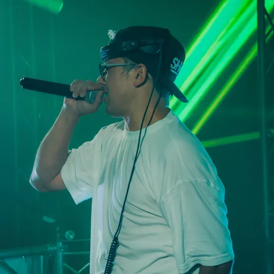 Ein Mann in einem weißen T-Shirt und einer schwarzen Mütze singt in ein Mikrofon, wobei ein grünes Licht den Hintergrund beleuchtet.