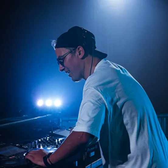 Ein Mann mit Brille und Kappe legt in einem schwach beleuchteten Raum mit blauen Lichtern auf. Er benutzt einen DJ-Mixer.