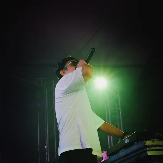 Ein Mann in einem weißen T-Shirt und Sonnenbrille singt in ein Mikrofon auf der Bühne, wobei ein grünes Licht den Hintergrund beleuchtet.
