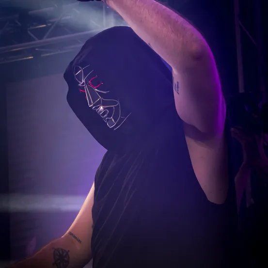 Eine Person in einem schwarzen Kapuzenpullover mit einer Maske auf der Bühne bei lila Beleuchtung performt.