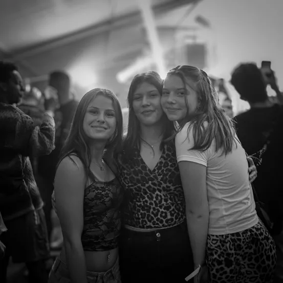 Drei junge Frauen lächeln und posieren für ein Foto in einem überfüllten, schwach beleuchteten Raum. Sie stehen eng beieinander und haben ihre Arme umeinander geschlungen.