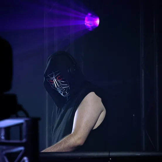 Eine maskierte Person in einem ärmellosen Shirt steht an einer DJ-Station unter einem violetten Scheinwerfer.