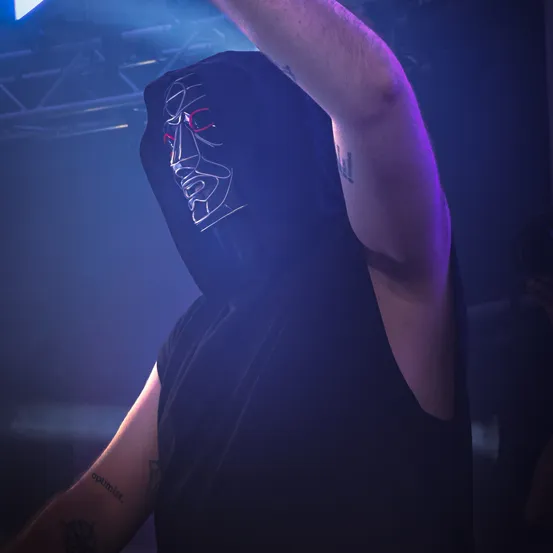 Eine Person mit Tätowierungen an den Armen trägt ein dunkles Kapuzenshirt mit Gesichtsdesign, das von roten Lichtern bei einem Konzert oder Event beleuchtet wird.