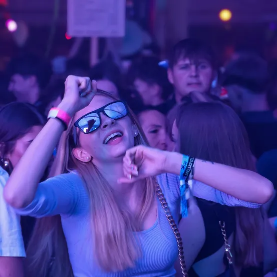 Eine junge Frau mit blonden Haaren tanzt bei einem Konzert mit einer Menge. Sie trägt eine Sonnenbrille und ein Armband. Der Veranstaltungsort ist mit lila Lichtern schwach beleuchtet.