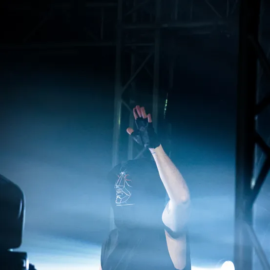Eine Person in einem schwarzen Outfit mit Maske, die ihre Hände unter blauem Licht in die Luft reckt.