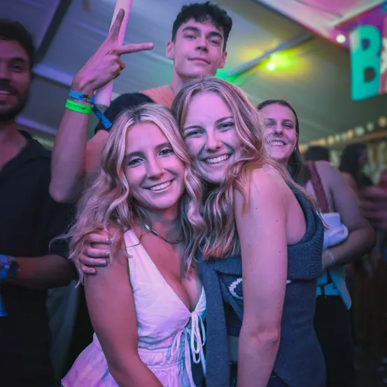 Zwei Frauen lächeln und posieren für ein Foto auf einer Party, mit Freunden dahinter, einer macht ein Friedenszeichen. Sie sind unter einem Zelt mit bunten Lichtern.