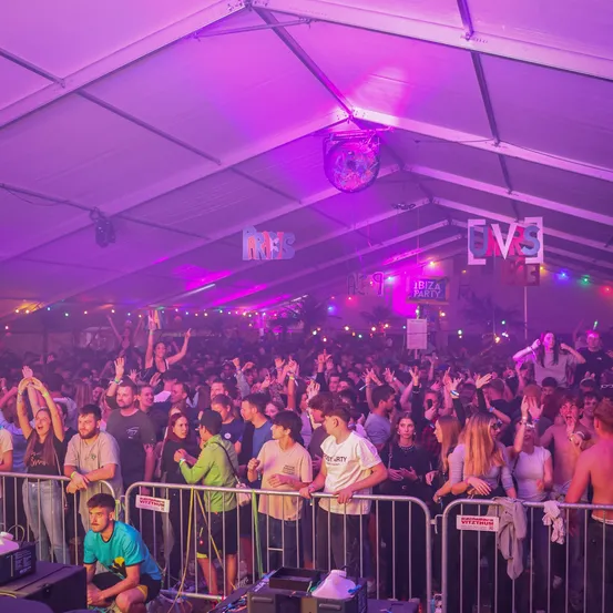 Eine Menschenmenge unter einem Zelt mit lila Beleuchtung, einer an der Decke hängenden Discokugel und Schildern mit der Aufschrift "UVS" und "IBIZA PARTY". Viele Menschen heben ihre Hände, und einige halten Schilder hoch.