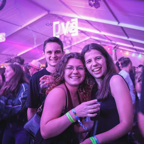 Drei Menschen lächeln und posieren für ein Foto bei einem Musikfestival unter einem lila Licht. Eine Frau hält ein Getränk und eine Kamera.