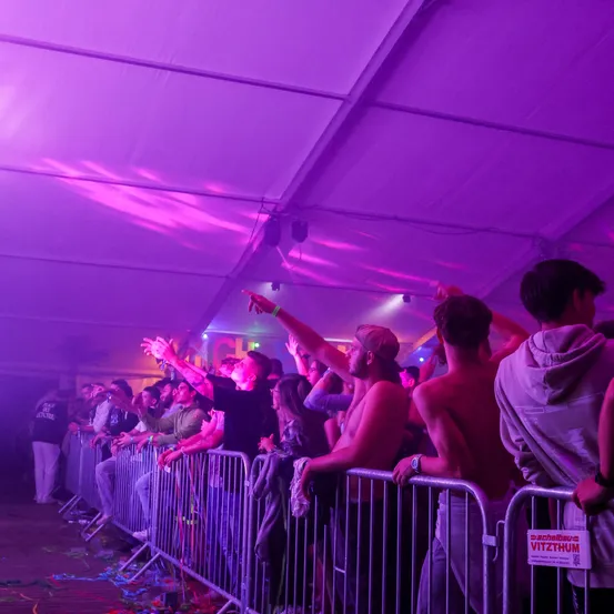 Eine Gruppe von Menschen steht hinter einer Metallbarriere, möglicherweise bei einem Konzert. Einige von ihnen heben ihre Hände. Der Bereich ist mit lila Lichtern beleuchtet. Die Person auf der linken Seite trägt eine Armbanduhr und eine Uhr. Die Metallbarriere hat ein Schild mit der Aufschrift 'VITZTHUM'. In der Ferne stehen auch Menschen.