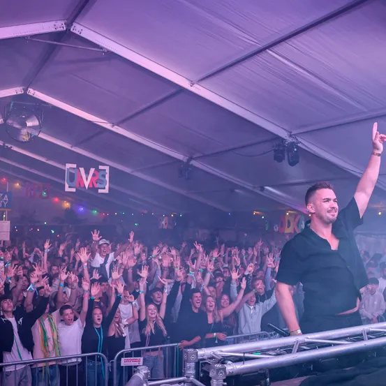 Ein DJ bei einem Musikfestival hebt seine Faust, während eine Menge jubelt, beleuchtet von bunten Lichtern und einem großen Zelt.