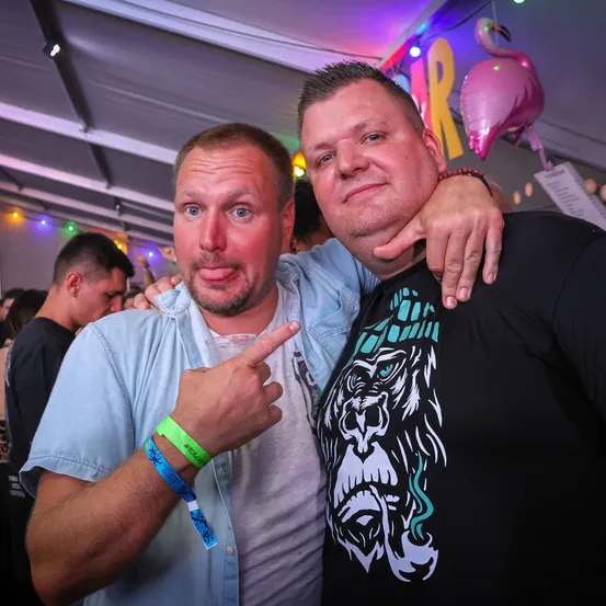 Zwei Männer posieren für ein Foto auf einer Party, einer zeigt auf die Kamera und sie haben Armbänder an den Handgelenken.