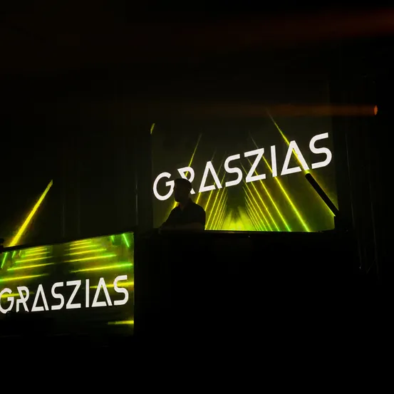 Eine Person steht hinter einem Podium, vor dem sich eine große Leinwand mit dem Wort GRASZIAS in fettweißen Buchstaben befindet. Grüne Laserlichter scheinen von unten auf die Leinwand.