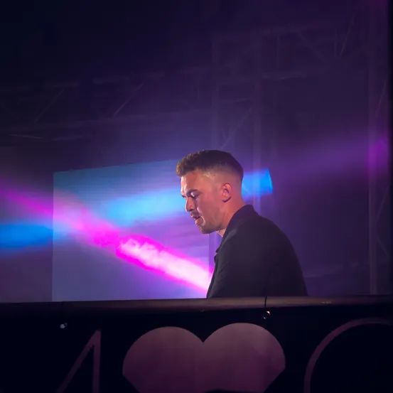 Ein Mann steht an einem DJ-Pult auf einer Bühne, beleuchtet von lila und blauen Lichtern. Ein großer Bildschirm ist hinter ihm.