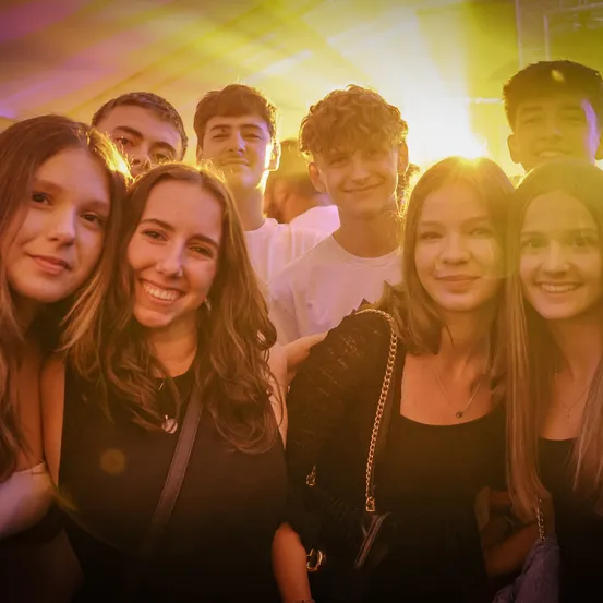 Eine Gruppe junger Menschen lächelt und posiert für ein Foto auf einer Party. Die Frauen tragen schwarze Oberteile und Halsketten, während die Männer weiße T-Shirts tragen. Heller Scheinwerfer beleuchten die Szene.