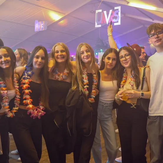 Eine Gruppe von Frauen und Männern lächelt für ein Foto in einem Partyraum. Sie tragen alle bunte Blumenkränze. Ein Mann hält ein Neonschild hoch. Der Raum ist schwach beleuchtet mit lila Lichtern.