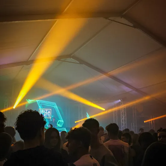 Eine Menschenmenge versammelt sich unter einem Zelt, das von hellen gelben Lichtern beleuchtet wird. Eine große Leinwand zeigt die Wörter 'Ibiza' und 'Party'. Ein DJ steht vor der Leinwand.