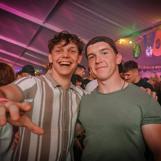 Zwei junge Männer lächeln und posieren für ein Foto auf einer Party mit bunten Lichtern.