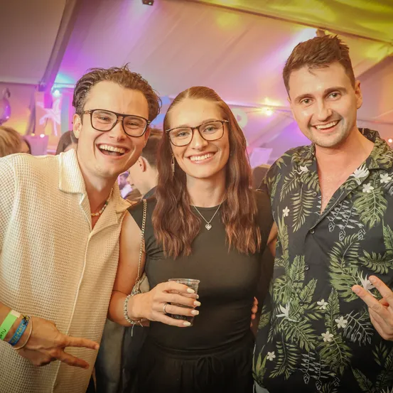 Drei Personen lächeln und posieren für ein Foto auf einer Party. Eine Frau hält ein Getränk, während ein Mann neben ihr zeigt. Hinter ihnen trägt ein Mann ein Blumenhemd.