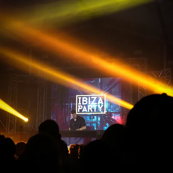 Ein DJ steht vor einem großen Bildschirm bei einer Ibiza-Party, mit hellen Bühnenlichtern, die die Menge beleuchten.