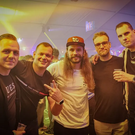 Fünf Männer posieren für ein Foto in einem schwach beleuchteten Raum. Ein Mann trägt eine Kappe und hat lange Haare. Der Mann zu seiner Linken macht ein Friedenszeichen.