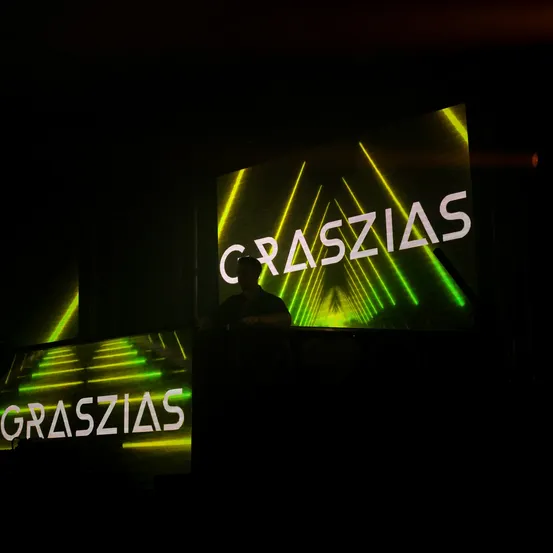 Ein Mann sitzt an einer DJ-Konsole mit zwei großen Bildschirmen hinter ihm, die das Wort 'Graszias' in Weiß auf grünem Hintergrund anzeigen.