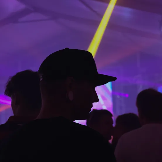 Menschen tanzen unter lila und gelbem Licht. Der Mann vorn trägt eine schwarze Kappe.