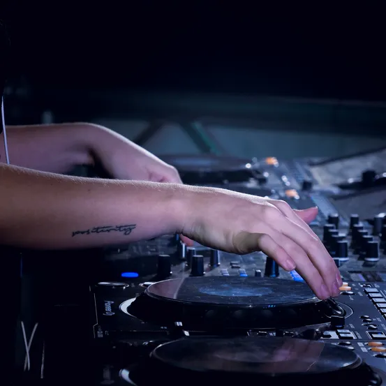 Eine Person mit einer Tätowierung am Arm legt auf und benutzt ihre Hände, um einen DJ-Mixer in einem schwach beleuchteten Raum zu steuern.