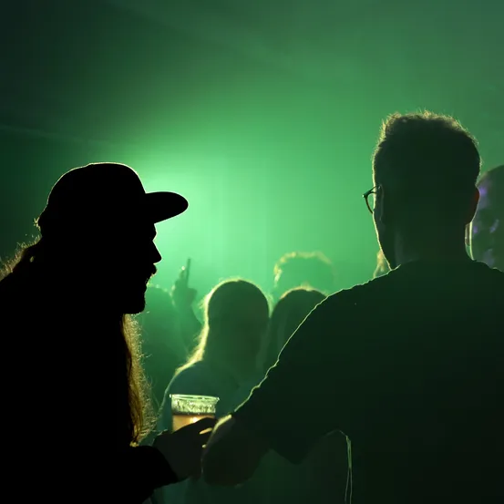 Die Silhouette eines Mannes mit einem Bierkrug, der einem anderen Mann in einem dunklen Raum mit grüner Beleuchtung gegenübersteht. Hinter ihnen stehen Menschen und heben ihre Hände.