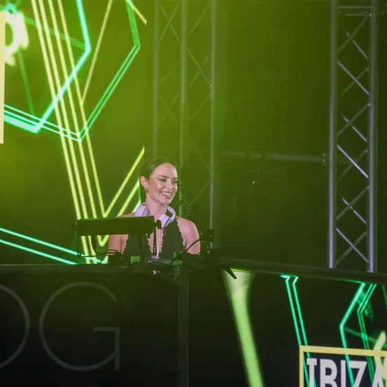 Eine Frau steht lächelnd an einem DJ-Pult unter einer grünen Lichtshow mit geometrischen Formen.