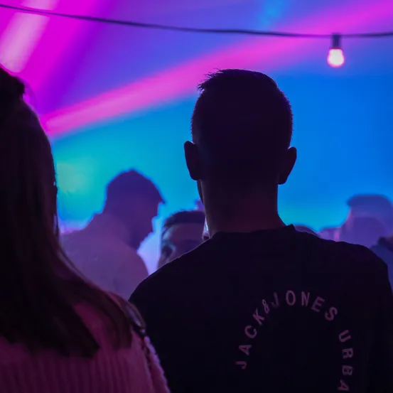 Eine Gruppe von Menschen befindet sich in einem Club mit lila und blauer Beleuchtung. Ein Mann in einem schwarzen T-Shirt mit Jack & Jones Urban schaut etwas an.