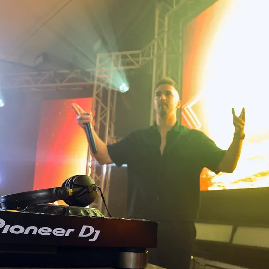 Ein Mann in einem schwarzen Shirt steht auf der Bühne, hält ein Mikrofon und trägt Kopfhörer. Vor ihm steht ein Pioneer DJ Controller. Hinter ihm zeigt eine große Leinwand einen Sonnenuntergang.