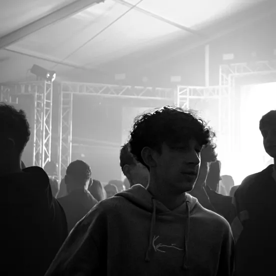 Ein Schwarz-Weiß-Foto zeigt eine Gruppe von Menschen in einem überfüllten Raum, möglicherweise bei einem Konzert. Ein Mann mit Kapuzenpullover und lockigem Haar steht in der Mitte, während andere im Hintergrund verschwommen sind.