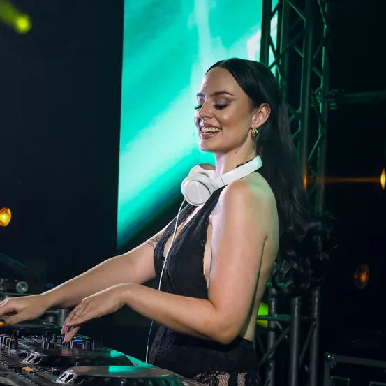 Eine Frau mit Kopfhörern steht an einem DJ-Mixer und lächelt in die Kamera. Im Hintergrund befindet sich eine grüne Projektionsleinwand und Bühnenbeleuchtung.