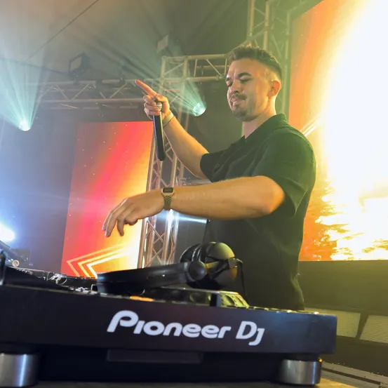 Ein DJ steht an einem Pioneer DJ-Turntable, zeigt mit einem Mikrofon auf das Publikum und steht unter einer farbenfrohen Bühnenbeleuchtung.