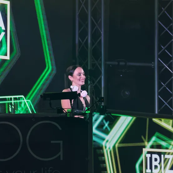 Eine lächelnde Frau steht auf einer DJ-Bühne mit Kopfhörern, grünen und gelben Lichtern im Hintergrund und einem Banner, das 'IBIZ' steht.