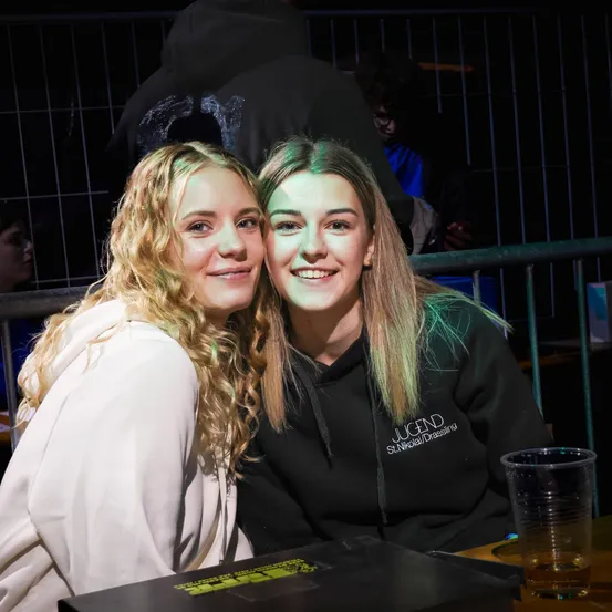 Zwei Frauen posieren für ein Foto mit einem Metallgitter und Getränken im Hintergrund, eine trägt eine weiße Kapuze, die andere eine schwarze mit 'JUGEND' darauf.