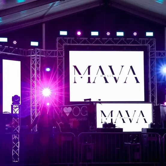 Eine Bühnenaufbau mit zwei großen Bildschirmen, die das 'MAVA MUSIC'-Logo unter lila Beleuchtung anzeigen, mit Scheinwerfern und Ausrüstung sichtbar.