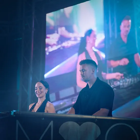 Ein Mann und eine Frau stehen lächelnd hinter einem DJ-Pult mit Herzsymbol. Dahinter zeigt eine große Leinwand einen DJ und zwei Personen, von denen eine ein Instrument spielt.