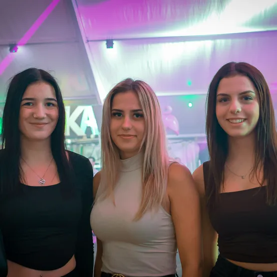 Drei junge Frauen stehen eng beieinander in einem neonbeleuchteten Raum und lächeln für ein Foto. Sie tragen verschiedenfarbige Oberteile, eine trägt ein schwarzes Crop-Top. Hinter ihnen steht ein Schild mit der Aufschrift 'KA'.
