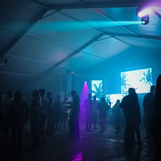 Eine Nachtszene mit Menschen in einem Tanzclub. Eine Bühne mit einem DJ in der Mitte, beleuchtet von blauen und lila Lichtern. Große Bildschirme mit dem Text 'IBIZA BEACH PARTY' auf ihnen.