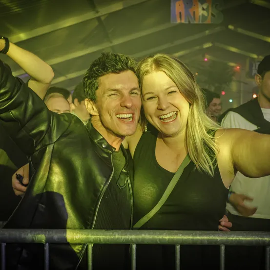 Ein Mann und eine Frau lächeln und posieren für ein Foto bei einem Konzert. Sie stehen nah an einer Metallbarriere.
