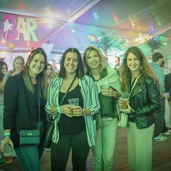 Vier Frauen stehen zusammen an einer Bar, lächeln und halten Getränke in den Händen. Sie sind von anderen Leuten umgeben. Über ihnen steht ein Schild mit der Aufschrift 'BAR'.