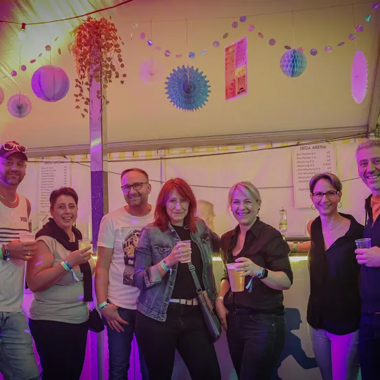 Eine Gruppe von Menschen in einem Partyzelt hält Getränke in der Hand und lächelt für ein Foto. Das Zelt ist mit bunten Papierlaternen und Luftballons dekoriert.