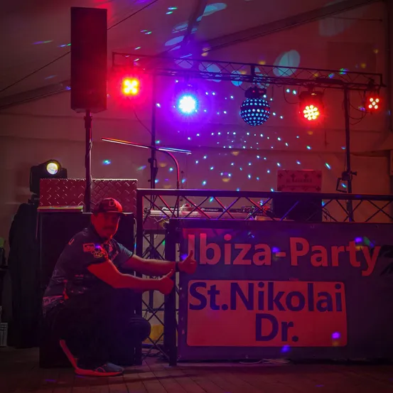 Ein Mann in einem grauen Shirt und einer Kappe justiert Ausrüstung auf einer Bühne mit bunten Lichtern. Die Bühne ist für eine Party vorbereitet mit einem Schild, das 'Ibiza-Party St.Nikolai Dr.' steht. Es sind Lautsprecher und eine DJ-Einrichtung sichtbar.