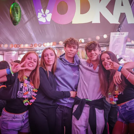 Fünf junge Leute posieren für ein Foto unter einem großen rosa und weißen 'VODKA'-Schild. Sie alle lächeln und scheinen eine gute Zeit zu haben.